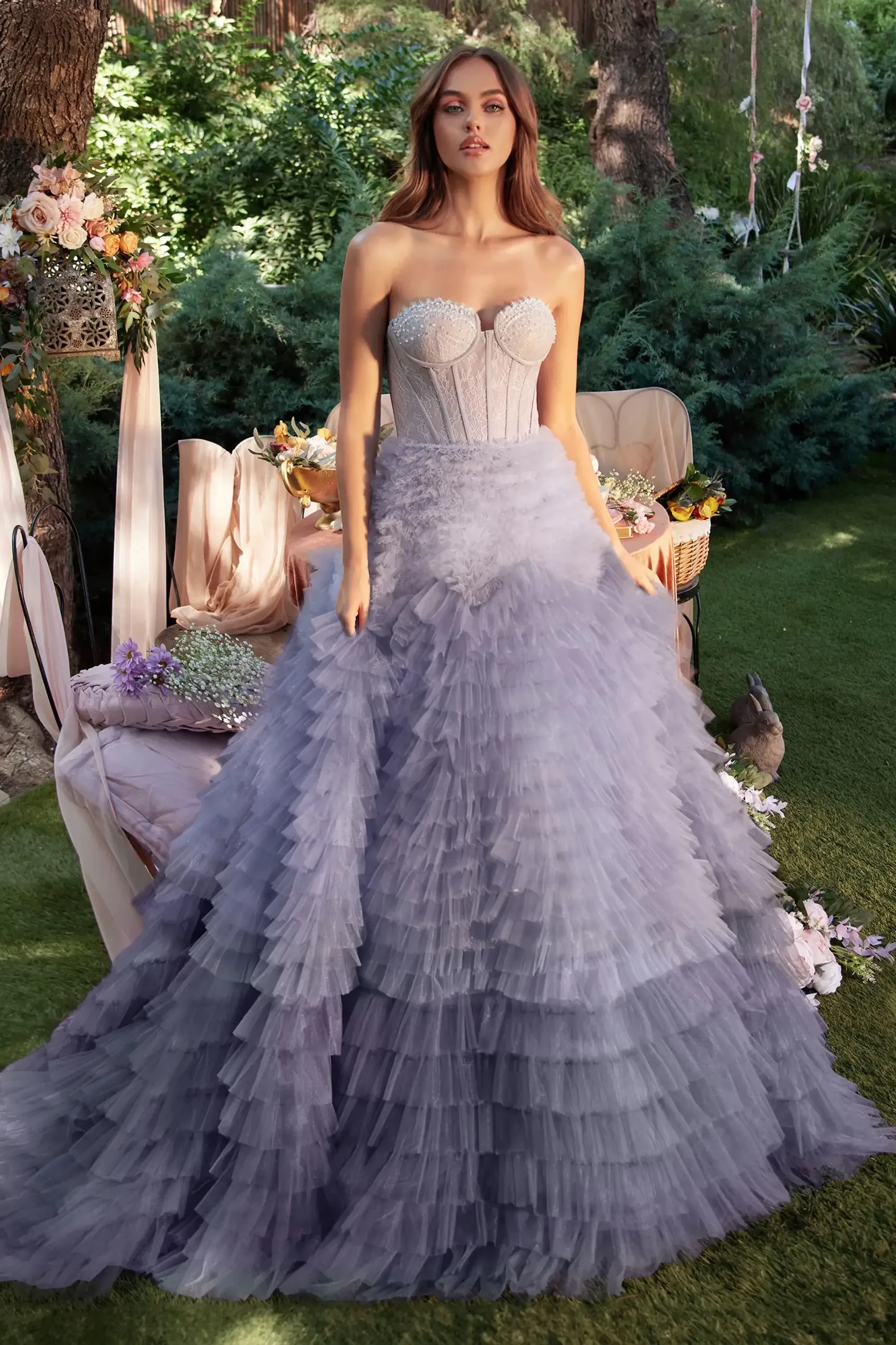 A woman stands outdoors in a flowing gray gown with ruffles and a structured bodice, surrounded by greenery and floral decorations.